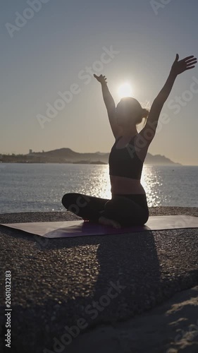 A person practices yoga on the beach. The sun rises, creating a glow behind them. The ocean waves gently lap at the shore. This is a time for focus and stretching.