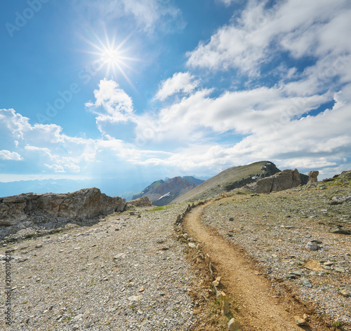 Scenic hiking trail winding through a rocky mountain ridge under a bright sun burst sky. Beautiful alpine landscape.