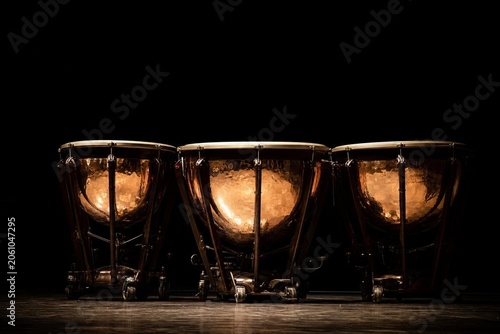 Three timpani drums with polished copper bowls reflecting light on a dark stage.