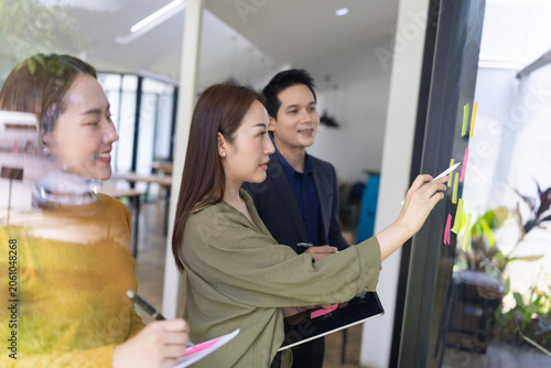 A group of young Asian business colleagues brainstorming and sharing ideas during a meeting, pointing at colorful sticky notes on a glass window in a modern office.