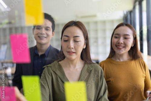 A group of young Asian business colleagues brainstorming and sharing ideas during a meeting, pointing at colorful sticky notes on a glass window in a modern office.