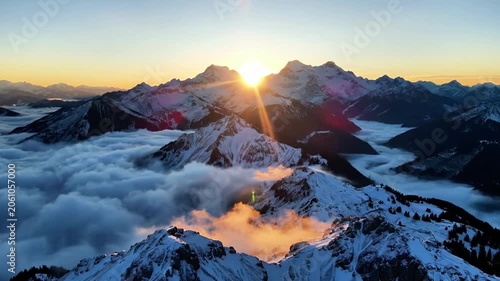 Aerial view of snow-capped mountains above clouds at sunrise with warm sunlight