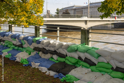 Green white and blue sandbags stacked as a long protective barrier wall on a riverbank promenade fence during an urban flood event. Emergency city defense and hazard protection