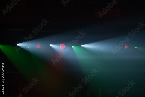 Stage spotlights pierce smoky air, colored beams sweep across empty stage, dramatic concert and theater lighting mood.