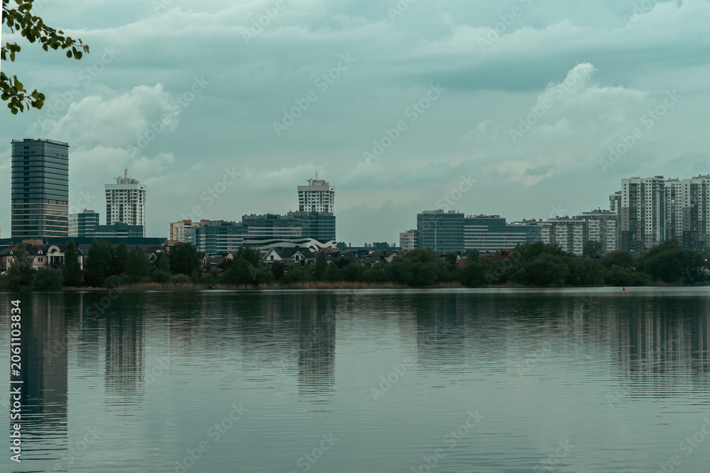 Fototapeta premium Modern Minsk residential district Lebyazhiy and business buildings reflected in calm water of Drozdy reservoir, urban waterfront landscape with green tree branches framing city skyline in Belarus