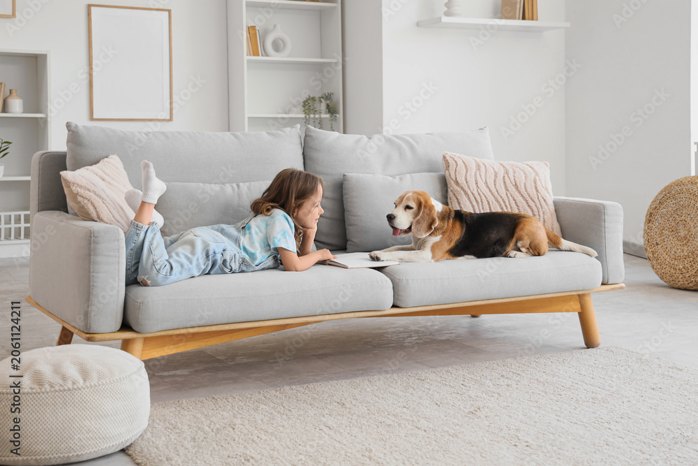 Naklejka premium Cute little girl with Beagle dog reading book on sofa in living room