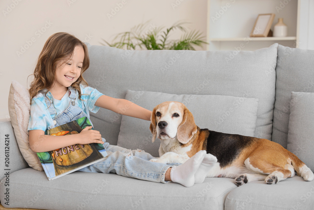 Naklejka premium Cute little girl with Beagle dog reading book on sofa in living room