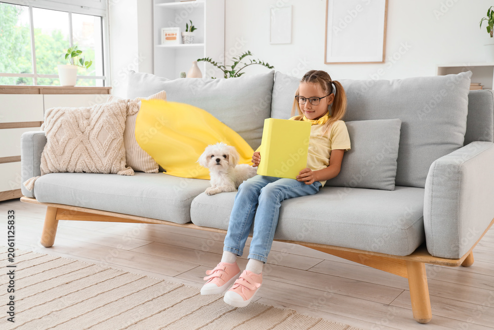 Naklejka premium Cute little happy girl in superhero costume with Maltese dog reading book on sofa at home