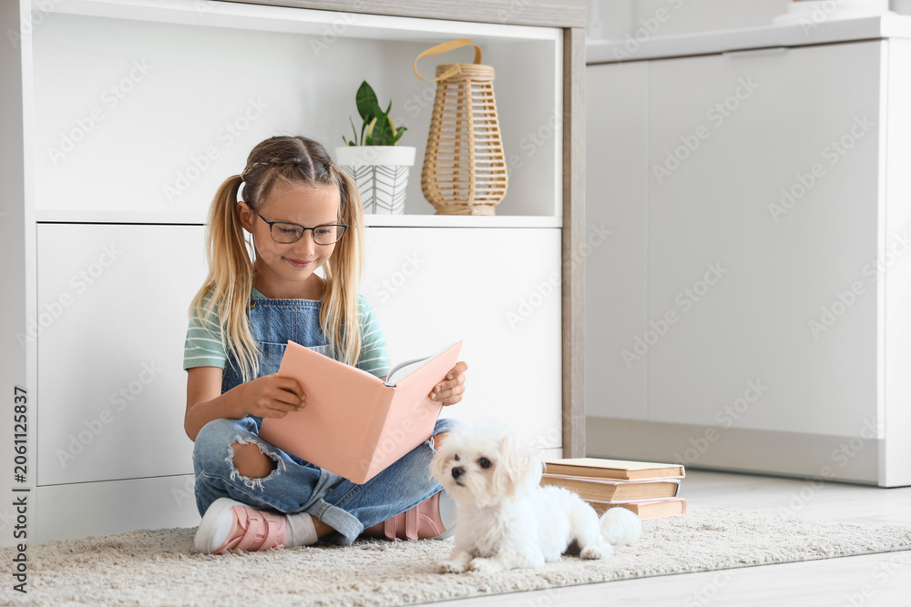 Naklejka premium Cute little happy girl with Maltese dog reading books on floor in kitchen