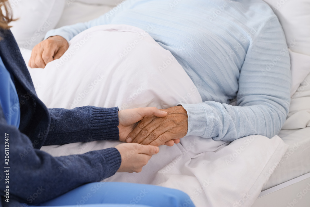 Fototapeta premium Caregiver with lying senior woman holding hands in bedroom, closeup. Care concept