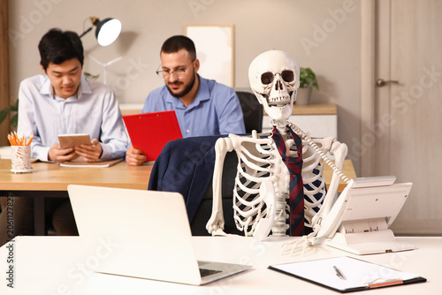 Human skeleton with telephone at table against male colleagues working in office