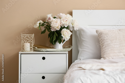 Burning candle and vase with peony flowers on table in bedroom