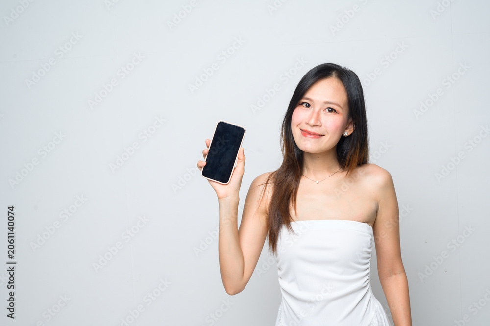 Naklejka premium A young woman in a white dress smiles while holding up a smartphone against a plain white background.