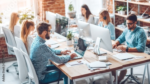 Multiple office workers collaborate and focus on tasks at shared wooden desks in a bright workspace.