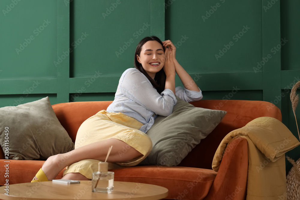 Fototapeta premium Young woman with cushions sitting on red sofa near green wall at home
