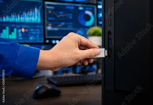 Cyber security analyst inserting USB drive into desktop computer, working in modern office environment with multiple monitors displaying data analytics, system monitoring, and digital interface.