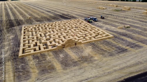 Intricate maze from hay bales stretches on agricultural field. Tractors near the entrance, seasonal farm activity aerial arc view
