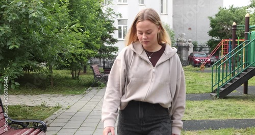Confident young woman with long blonde hair walking along a park path, approaching the camera, turning around with a bright smile, and continuing her stroll on a pleasant summer day