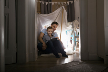 Father and son sitting in self-made tent at home in the evening using digital tablet