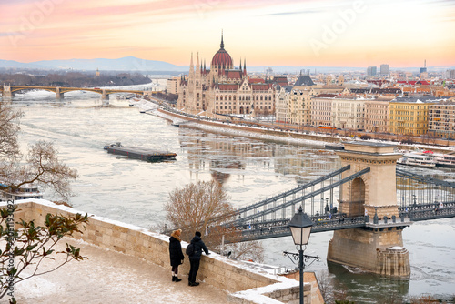 Tourists looking at winter panorama of Budapest with Hungarian Parliament and Chain Bridge over frozen Danube river at sunset, Hungary