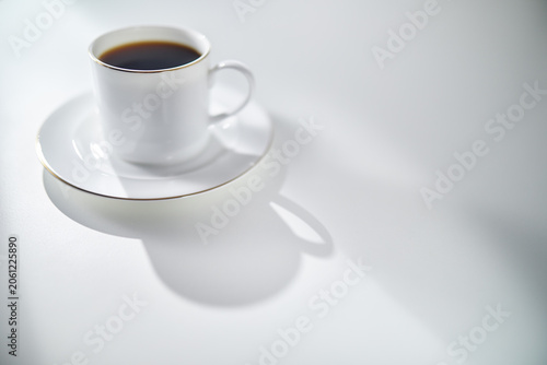 Elegant White Coffee Cup with Gold Rim and Saucer on a White Table in Soft Morning Light with Copy Space. Luxury lifestyle concept. Shallow depth of field.