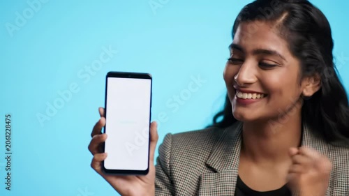 Diverse young professional in business-casual blazer enthusiastically reacts to a smartphone, smiling, nodding, and fist-pumping, then looks up, on bright pastel background. Positive breakthrough