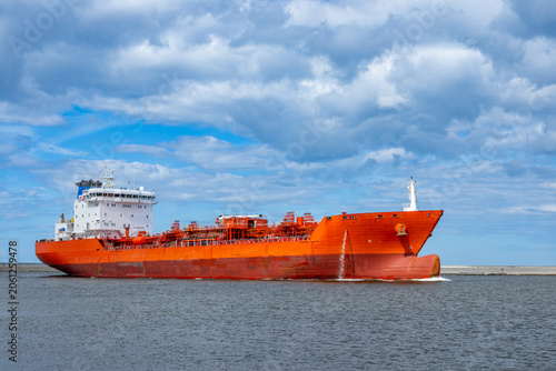 Large orange cargo ship sailing on calm sea under dramatic cloudy sky near harbor entrance