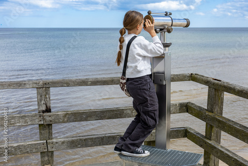Child looking through telescope on wooden seaside pier with calm sea horizon and cloudy sky