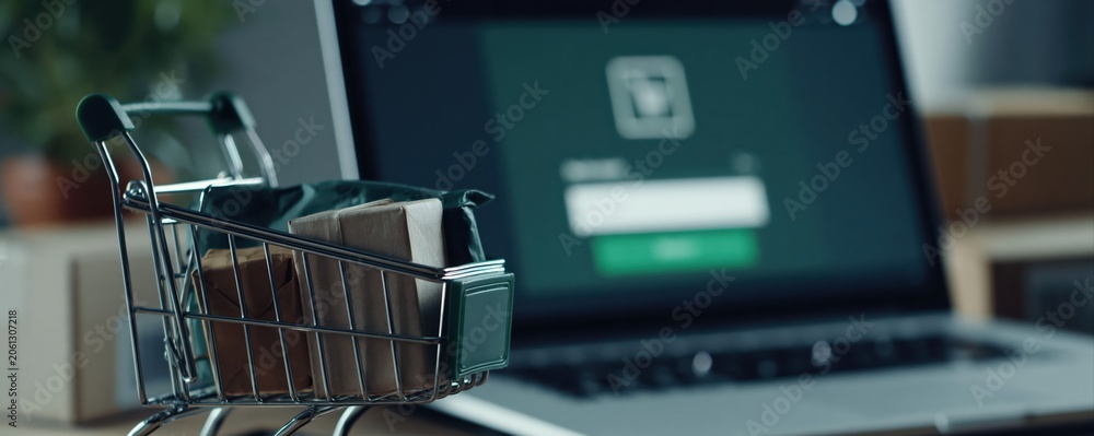 Fototapeta premium Premium commercial photo of a laptop with a checkout interface and a mini shopping trolley filled with parcels. Perfect visual for ecommerce, online shopping, and retail business themes.