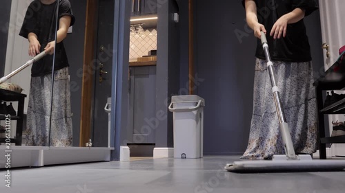 A teenager mops a gray tile entryway floor beside a mirrored closet and gray bucket during a household chore.
