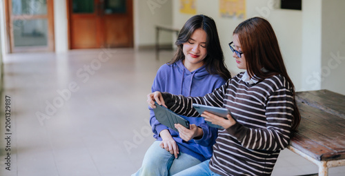 Two high school teenagers sitting together in school hallway using digital tablets during peer collaboration and social media interaction