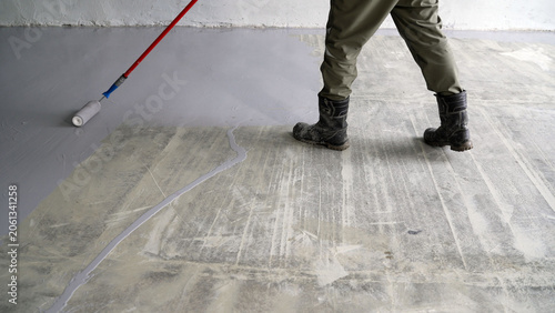 Construction worker applying gray floor coating to concrete surface. Close up of legs and boots during renovation. Industrial flooring and maintenance