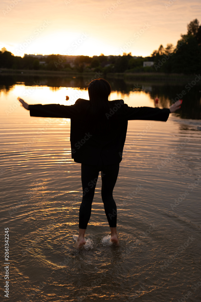 Fototapeta premium silhouetted figure wading barefoot into a shallow lake at golden sunset, arms extended in a peaceful, contemplative pose; warm orange-pink sky reflected in rippling water, dark treeline on the horizon