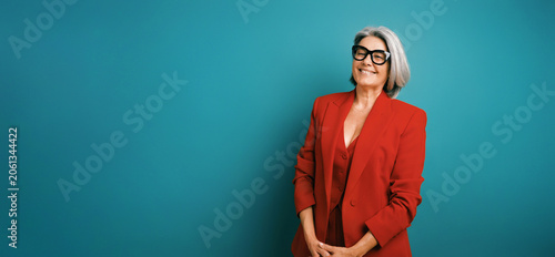 Senior businesswoman in red blazer and stylish glasses expressing confidence and joy, standing on a teal studio backdrop