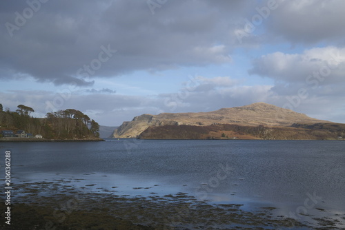 Early evening coastal landscape of Isle of Skye, Portree, Scotland, with deep blue sea and dramatic clouds above mountains and tiny red boat in distance