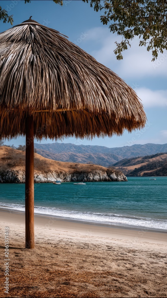 Fototapeta premium Low angle view of a large thatched umbrella on a beach with turquoise ocean and distant mountains under a partly cloudy sky