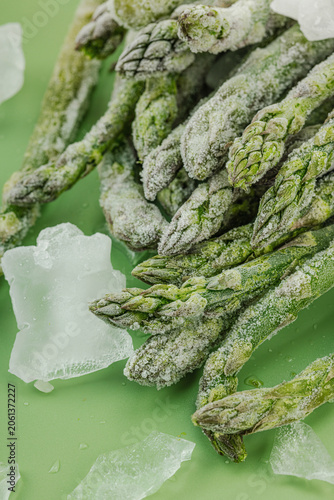 Frozen green asparagus spears covered in hoarfrost with ice cubes on a green background