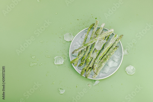 Frozen green asparagus spears covered in hoarfrost with ice cubes on a green background