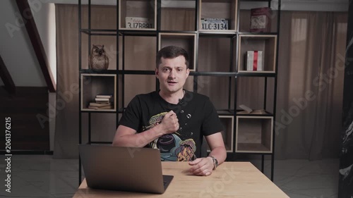 Man in t-shirt in front of laptop talking and punch in the chest. Shelf with books on background. Interview concept.