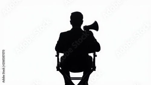 Rear view of an Asian man in a black director's chair, holding a megaphone against a white backdrop. Silhouette image.