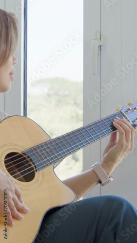Vertical video: Panning camera revealing woman singing, strumming guitar by window, practicing song