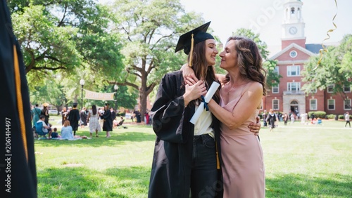 Mother hugs graduate daughter wearing black cap and gown with diploma on university grounds.