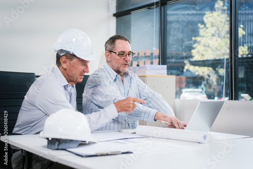 Two senior men review building plans in an office, using a laptop and documents. Safety helmets and blueprints suggest engineering, construction planning, teamwork and project discussion.