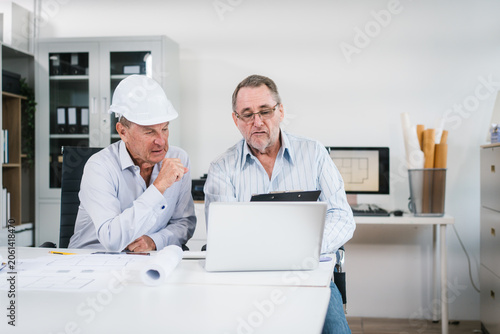 Two senior men review building plans in an office, using a laptop and documents. Safety helmets and blueprints suggest engineering, construction planning, teamwork and project discussion.