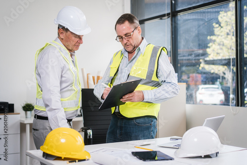Two construction professionals in safety vests and hard hats review plans on a clipboard in a bright office, with helmets and documents on the table.