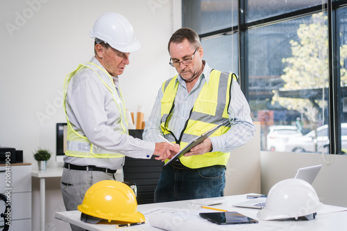 Two construction professionals in safety vests and hard hats review plans on a clipboard in a bright office, with helmets and documents on the table.
