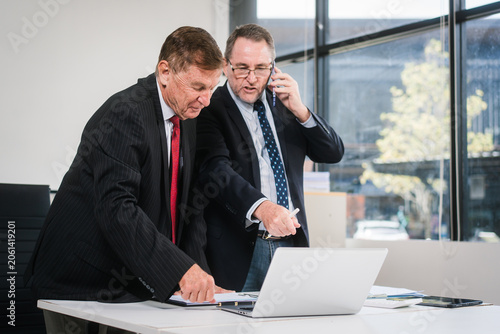 Two businessmen in suits discuss documents and a laptop in a bright modern office, with one man talking on the phone during a focused business meeting.