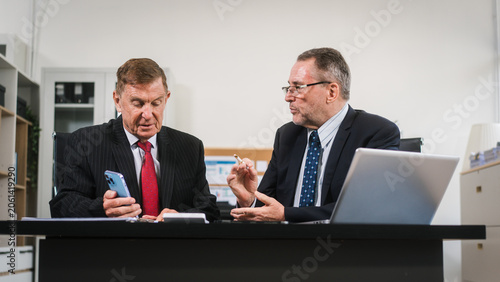Two senior businessmen in suits working together at an office desk, reviewing documents and a smartphone beside a laptop in a modern professional setting.