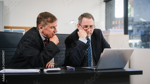 Two business men in suits sit at an office desk looking stressed and frustrated during a difficult meeting, with a laptop and paperwork in front of them.