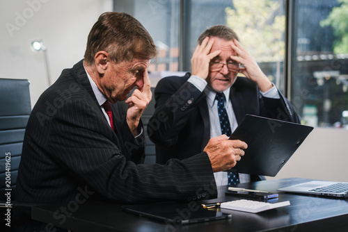Two business men in suits sit at an office desk looking stressed and frustrated during a difficult meeting, with a laptop and paperwork in front of them.
